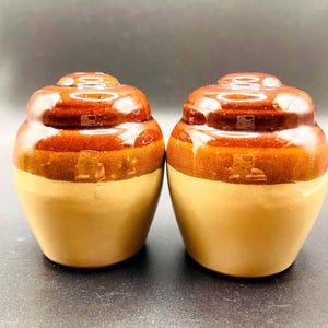 Könnte beinhalten: Zwei Keramikgläser mit Deckel. Die Gläser haben ein zweifarbiges Design mit einem hellbraunen Sockel und einem glänzenden braunen Oberteil. Die Deckel sind abgerundet und haben eine kleine, quadratische Vertiefung. Die Gläser sind wahrscheinlich für die Küchenaufbewahrung.