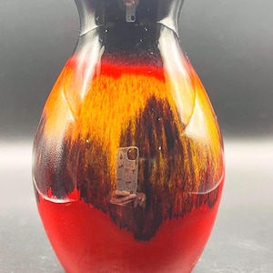 Puede incluir: Un jarrón decorativo de cerámica con acabado brillante. El jarrón presenta un degradado de color, que va desde una base roja intensa hasta una sección media naranja y marrón ardiente, culminando en un borde negro. El jarrón tiene un cuerpo bulboso y un cuello estrecho.