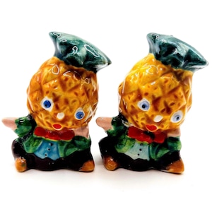 May include: A pair of vintage ceramic pineapple-shaped salt and pepper shakers. Each shaker is painted with a yellow pineapple body, blue eyes, a red bow tie, and a green hat and jacket. The shakers are approximately 7.6 cm tall.