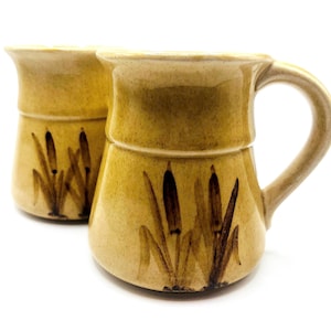 Puede incluir: Dos tazas de cerámica con un esmalte color beige y un diseño de junco pintado a mano en marrón. Cada taza tiene un asa y un borde ligeramente acampanado. Las tazas son adecuadas para bebidas frías o calientes.