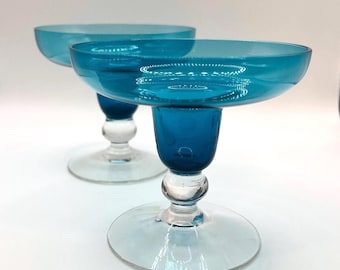 May include: Two matching coupe glasses featuring wide, shallow teal blue bowls. Each glass has a short, thick teal blue stem connected to a clear glass base by a small, rounded clear glass knob. The design is smooth, polished, and translucent.