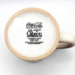 May include: Cream-colored ceramic mug with a Coca-Cola logo and branding information. The bottom of the mug features the Coca-Cola logo, the Gibson brand, and text indicating it is dishwasher, oven, and microwave safe.