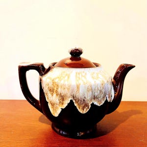 May include: A vintage brown ceramic teapot with a lid and handle. The teapot has a unique glaze design with a white and tan drip pattern over the dark brown base. The teapot is sitting on a wooden surface.