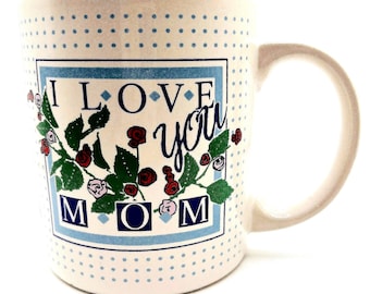 May include: White ceramic mug with a blue dotted pattern and a handle. The mug features a rectangular design with the words "I LOVE YOU MOM" surrounded by floral illustrations of red and pink roses and green leaves.