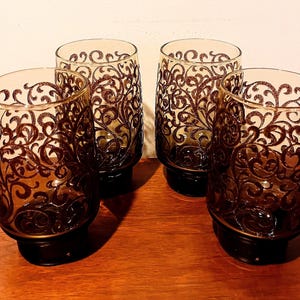 Può includere: Quattro bicchieri vintage con un motivo a vortice marrone ornato. I bicchieri hanno una base marrone scuro e una parte superiore trasparente. I bicchieri sono disposti su una superficie di legno.