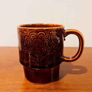 Könnte beinhalten: Eine braune Keramiktasse mit floralem Muster. Die Tasse hat einen dunkelbraunen Boden und einen helleren braunen oberen Teil mit Henkel. Das florale Design ist auf der Oberfläche der Tasse geprägt.