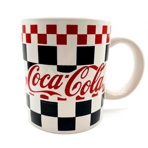 May include: White ceramic mug with a red, black, and white checkered pattern. The Coca-Cola logo is prominently displayed in red script across the front of the mug. The mug has a curved handle and a glossy finish.
