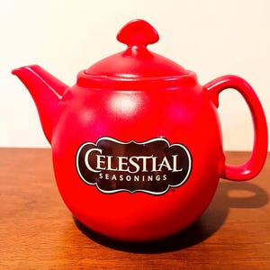 May include: A bright red ceramic teapot with a lid and handle. The teapot has the "Celestial Seasonings" logo in a black and white oval. The teapot is designed for brewing tea.
