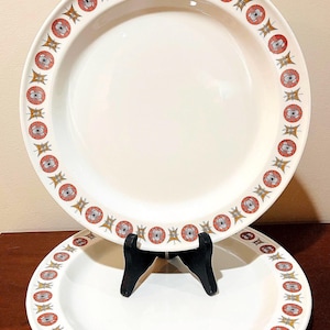 Peut inclure: Deux assiettes en céramique blanche avec une bordure décorative présentant un motif répétitif de motifs floraux rouges, oranges et jaunes. Une assiette est sur un support noir, l'autre au premier plan.