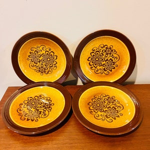 May include: Four vintage ceramic plates with a floral design. The plates have a brown rim and a yellow centre with a brown floral pattern. The plates are arranged on a wooden surface.