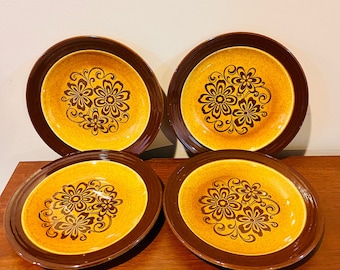May include: Four vintage ceramic plates with a floral design. The plates have a brown rim and a yellow center with a brown floral pattern. The plates are arranged on a wooden surface.