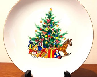 May include: White decorative plate with a gold rim featuring a Christmas tree design. The tree is decorated with ornaments, bows, and candy canes. Presents and a rocking horse are at the base of the tree.