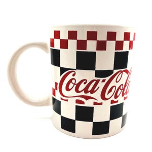 Może przedstawiać: Biały ceramiczny kubek Coca-Cola z czerwono-czarno-białym wzorem w szachownicę. Logo Coca-Cola jest w czerwonym skrypcie. Kubek ma biały uchwyt i błyszczące wykończenie.