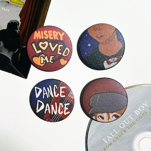 Puede incluir: Cuatro botones redondos con pasador con diferentes diseños. El primer botón es rojo con texto blanco que dice "Misery Loved Me" con dos corazones. El segundo botón es azul con una ilustración en blanco y negro de una cara. El tercer botón es rojo y azul con texto blanco que dice "Dance Dance". El cuarto botón es rojo con una ilustración en blanco y negro de una cara con un sombrero.