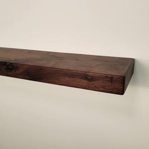 May include: A dark brown, rectangular floating shelf made of wood. The shelf is mounted on a white wall and has a natural wood grain pattern. The shelf is approximately 30 inches long.