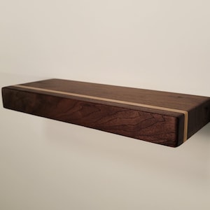 Puede incluir: Estante flotante rectangular de madera marrón oscuro con una franja clara. Mide aproximadamente 60 cm de largo y está montado en una pared blanca. Se aprecia la veta de la madera.
