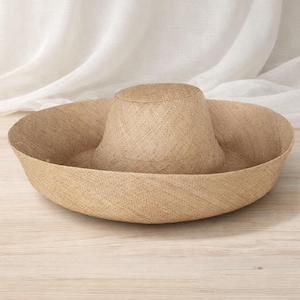 May include: A natural-coloured straw hat with a wide brim and a rounded crown. The hat features a woven texture and a broad, gently sloping brim. The hat is designed for sun protection. The hat is on a light-coloured wooden surface.