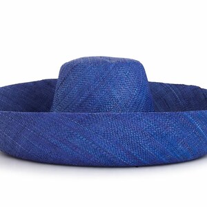 May include: A wide-brimmed hat crafted from woven straw in a vibrant royal blue colour. The hat features a rounded crown and a broad, slightly upturned brim with a visible diagonal weave pattern throughout the entire structure of the accessory.
