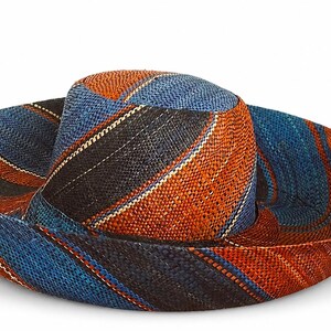 May include: A wide-brimmed woven hat featuring a diagonal striped pattern in shades of deep blue, burnt orange, and black. The hat has a rounded crown and a flexible, slightly upturned brim, constructed from a textured, straw-like material.