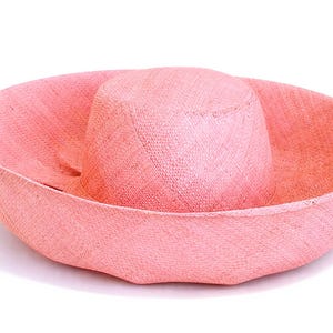 May include: A wide-brimmed hat crafted from woven straw material in a solid light pink colour. The hat features a rounded crown and a broad, slightly upturned brim with a consistent textured weave pattern throughout the entire surface of the object.