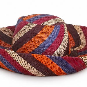 May include: A wide-brimmed hat with a colourful striped pattern. The hat features a combination of brown, orange, blue, red, and cream-coloured stripes. The hat's crown is cylindrical, and the brim is wide and slightly curved.