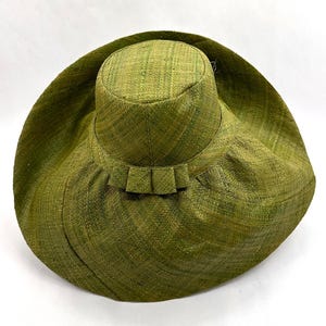 May include: A wide-brimmed hat crafted from woven green straw material. The hat features a tall, cylindrical crown and a broad, flexible brim. A decorative band with three small, folded rectangular pleats encircles the base of the crown.