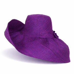 May include: A vibrant purple wide-brimmed sun hat crafted from a woven straw-like material. The hat features a rounded crown and a large, flexible brim that curves downward. A small, matching purple fabric bow is attached to the side of the crown.