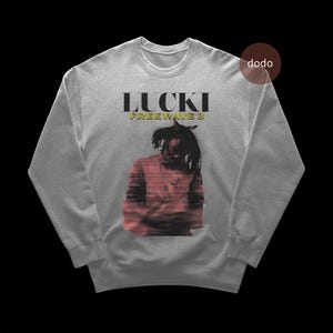以下が含まれることがあります： 黒と金の「LUCKI FREEWAVE 3」の文字が入ったグレーのスウェットシャツ。スウェットシャツには、赤と黒のデザインのドレッドヘアの人のグラフィックがあります。