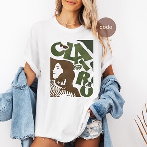 May include: A white short-sleeve t-shirt featuring a graphic print with the text CLAIRO in large olive green letters. The design includes stylized brown and olive green silhouettes of a face and eyes, layered over a white background.