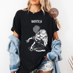 Könnte beinhalten: Schwarzes T-Shirt mit dem Wort "BONES" in weißen gotischen Buchstaben über einer Schwarz-Weiß-Illustration eines Skeletts mit Mütze. Das Skelett hockt.
