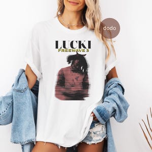 以下が含まれることがあります： 「LUCKI FREEWAVE 3」の文字と人物のグラフィックがプリントされた白いTシャツ。グラフィックは赤、黒、白の色合いです。Tシャツは半袖で、クルーネックです。