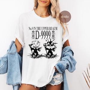Puede incluir: Camiseta blanca con un diseño gráfico negro. El diseño incluye el texto "No.9 IN THE UPPER REALM D-9999", calaveras y dos figuras de monstruos. La camiseta es de estilo casual.
