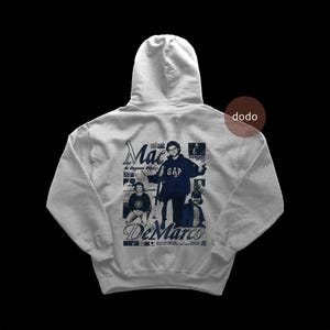 May include: A light gray hoodie featuring a graphic design with the text "Mac DeMarco" in a dark blue font. The design includes images of the musician and text that reads "the Peppermill Playhouse". The hoodie has a hood and a kangaroo pocket.