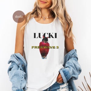 以下が含まれることがあります： 「LUCKI」の文字と赤と黒のグラフィック、そして金色の「FREEWAVE 3」の文字がプリントされた白いタンクトップ。ノースリーブで、柔らかい素材で作られています。