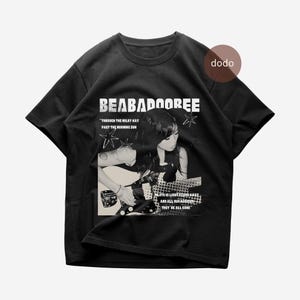 Op de afbeelding: Zwart T-shirt met de bandnaam "BEABADOOREE" in het wit. Het ontwerp bevat een zwart-wit afbeelding van een persoon die gitaar speelt, met songteksten en sterrenafbeeldingen.