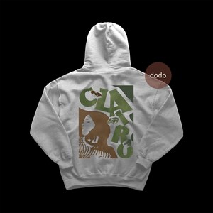 May include: A light gray hooded sweatshirt featuring a graphic print on the back. The design includes the text CLAIRO in large olive green letters, layered with brown silhouettes of a person and a detailed eye, all set against a solid background.