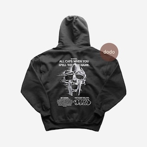 Könnte beinhalten: Ein anthrazitfarbener Kapuzenpullover mit weißer Grafik auf der Rückseite. Das Design zeigt eine metallische Maskenillustration, den Text ALL CAPS WHEN YOU SPELL THE MAN NAME, MF DOOM, THE ILLEST VILLAIN und ein DOOM-Logo im Graffiti-Stil.