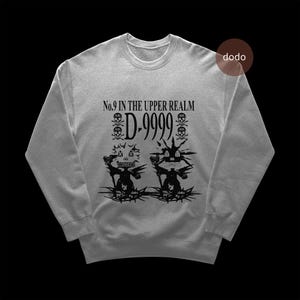 Puede incluir: Sudadera gris claro con cuello redondo. Presenta un diseño negro con el texto "No. 9 in the Upper Realm" y "D-9999", además de calaveras y elementos puntiagudos. El diseño es estilizado.