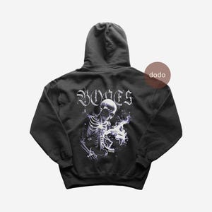 Könnte beinhalten: Schwarzer Kapuzenpullover mit einem Skelett-Motiv, das einen Globus mit Flammen hält. Das Wort "BONES" ist in gotischer Schrift auf dem oberen Rücken aufgedruckt. Der Hoodie hat eine Kapuze mit Kordelzug und eine Kängurutasche.