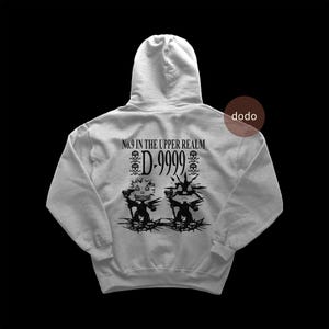 Puede incluir: Una sudadera con capucha gris claro con texto e ilustraciones en negro en la espalda. El texto dice "No.9 in the Upper Realm" encima de "D-9999" y dos símbolos de calaveras. Debajo hay dos figuras estilizadas.