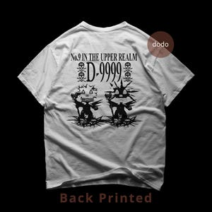 Puede incluir: Camiseta blanca con estampado gráfico negro en la espalda. El diseño incluye el texto "No.9 IN THE UPPER REALM" encima de "D-9999" y dos símbolos de calavera y huesos cruzados. Debajo, dos figuras estilizadas con detalles puntiagudos.