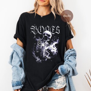 Könnte beinhalten: Schwarzes T-Shirt mit einem Skelett-Motiv, das einen Globus hält, mit lila und weißen Akzenten. Das Wort "BONES" ist in einer gotischen Schriftart über dem Skelett aufgedruckt. Das Wort "BLUNT" ist auf dem Globus aufgedruckt.
