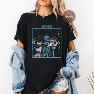 Könnte beinhalten: Schwarzes T-Shirt mit einem blau getönten Grafik eines Skeletts in einer Friedhofsszene, eingerahmt in einem blauen Quadrat. Das Wort "BONES" ist in Blau über der Grafik gedruckt. Das T-Shirt ist ein lässiger Stil.