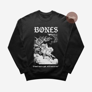 Könnte beinhalten: Schwarzes Sweatshirt mit dem Wort "BONES" in gotischer Schrift. Die Grafik zeigt eine detaillierte Schwarz-Weiß-Illustration von Figuren und Wolken. Das Zitat "I don't need a god, all I need is me" ist darunter gedruckt.