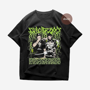May include: Black t-shirt with a neon green graphic of the text "SUICIDEBOYS", two figures, and "Greyday 2023". The design incorporates lightning bolt motifs and additional text at the bottom, all in a bold, eye-catching style.