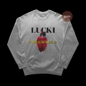 以下が含まれることがあります： グレーのスウェットシャツ。黒で「LUCKI」の文字が入り、赤い衣装を着た人のグラフィックが上にあります。「FREEWAVE 3」の文字は黄色のグラフィックの下にあります。スウェットシャツはクルーネックです。