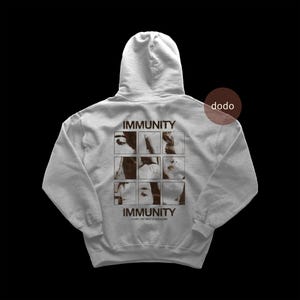 May include: A light gray hooded sweatshirt featuring a dark brown graphic on the back. The design includes the word IMMUNITY above and below a three-by-three grid of cropped photographic images, with the text CLAIRO THE DEBUT STUDIO ALBUM below.