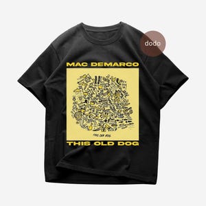 May include: A black short-sleeve t-shirt featuring a yellow square graphic on the chest. The graphic contains black line-art doodles and the text MAC DEMARCO and THIS OLD DOG in yellow capital letters. A small brown circle with the text dodo is visible.
