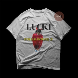 May include: A light gray t-shirt featuring the word "LUCKI" in black block letters above a graphic of a person in a red outfit. The words "FREEWAVE 3" are in gold block letters below the graphic.