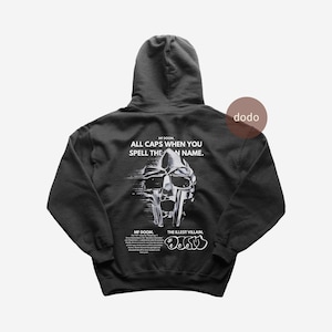 Puede incluir: Sudadera con capucha negra con un gráfico de una máscara y el texto "MF DOOM. ALL CAPS WHEN YOU SPELL THE NAME." La parte trasera de la sudadera también tiene el texto "THE ILLEST VILLAIN." y el logo "dodo".